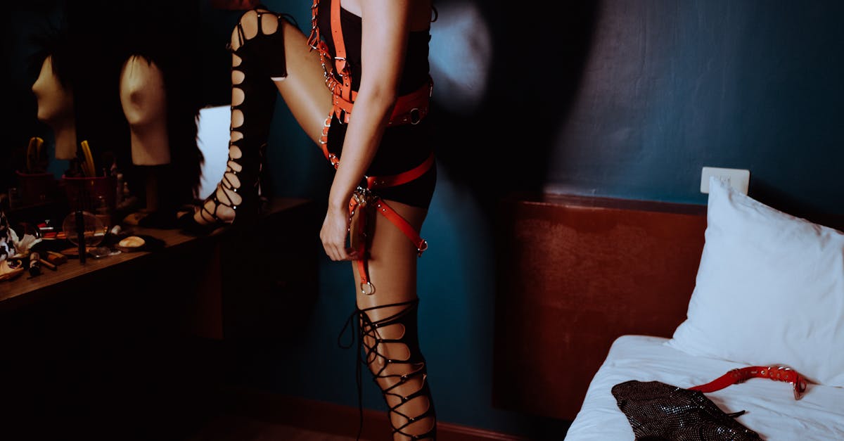 Side view of crop unrecognizable transsexual male in eccentric BDSM harness and bodysuit standing with leg on mirror table in bedroom