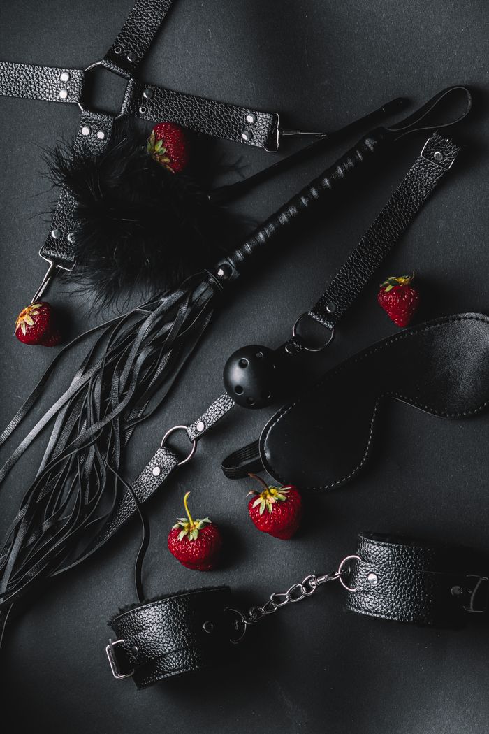 A stylish flat lay of leather BDSM accessories with fresh strawberries on a dark background.